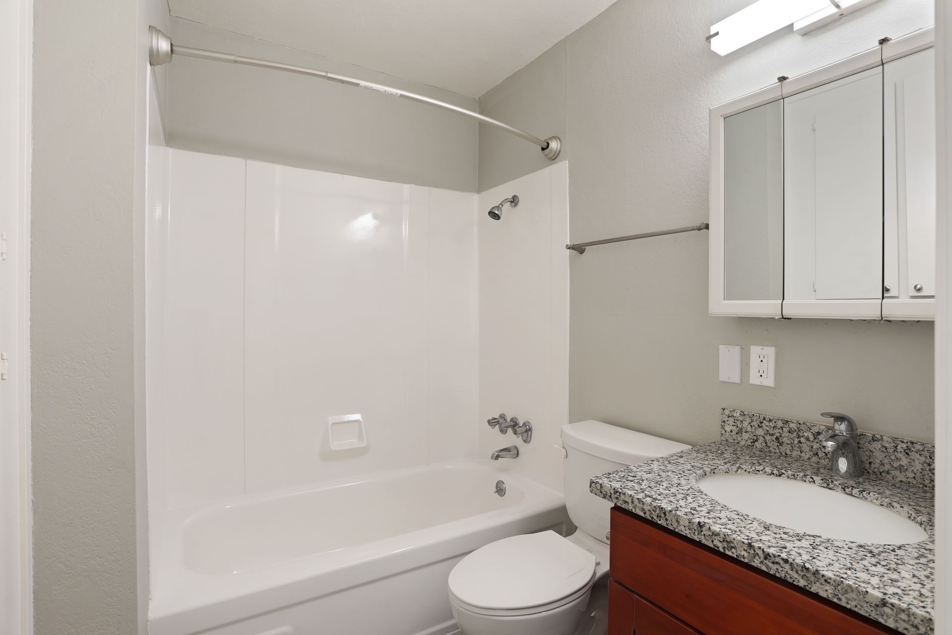 Bright bathroom with white tub-shower, toilet, granite vanity, and mirrored cabinet.