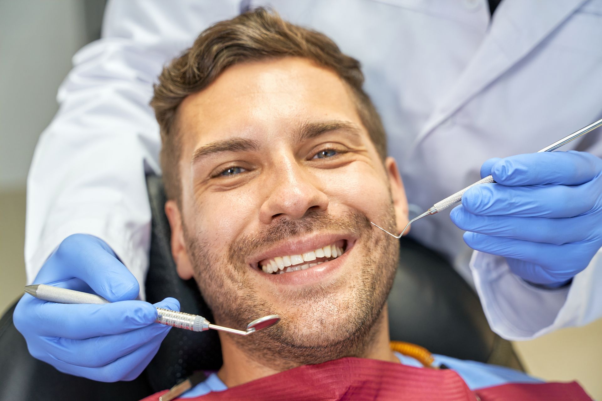 Man smiling in a dentist chair, dentist holding tools, blue gloves, white coat.