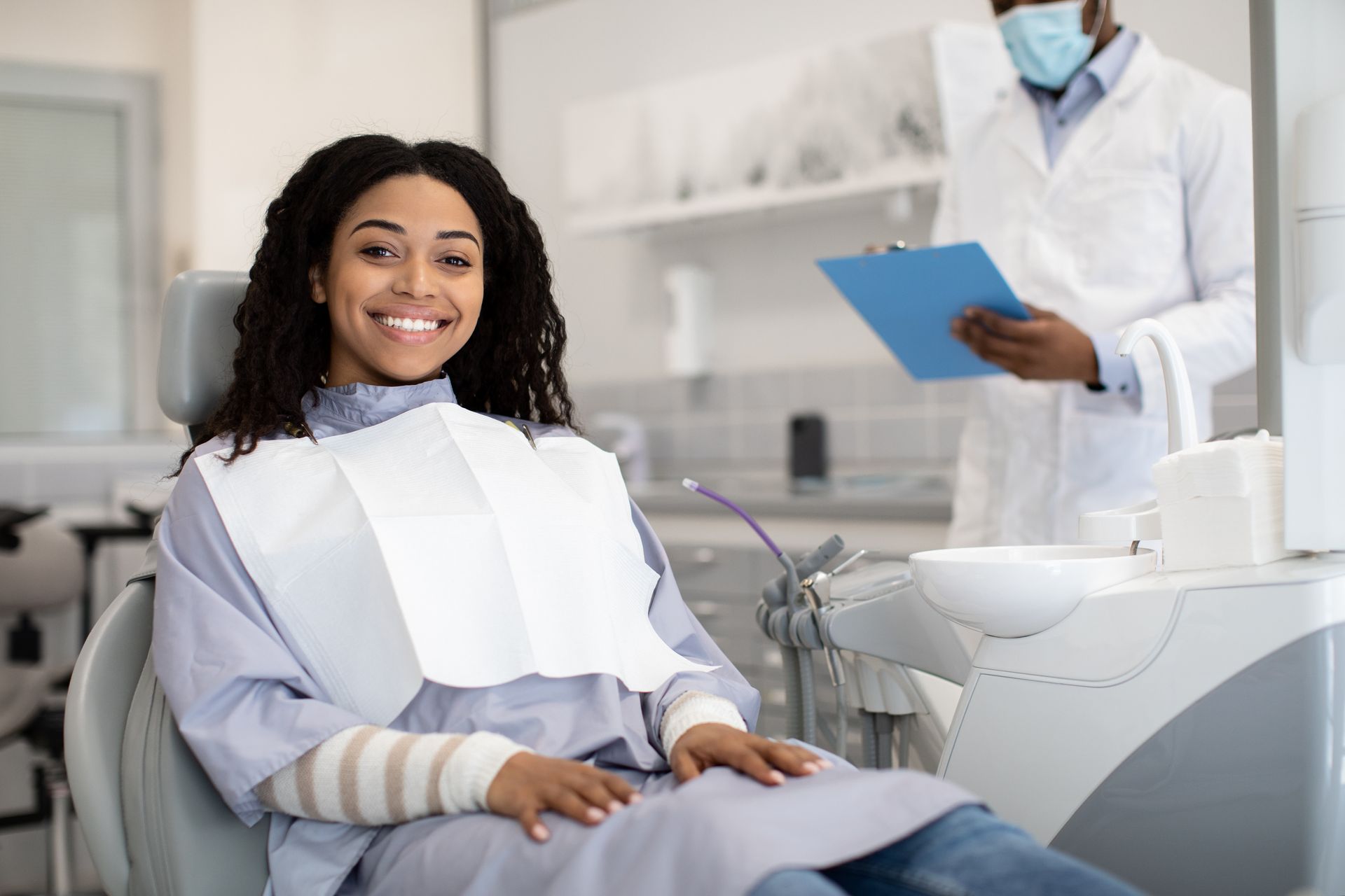Woman in dentist chair smiles; dentist in mask and coat holds clipboard.