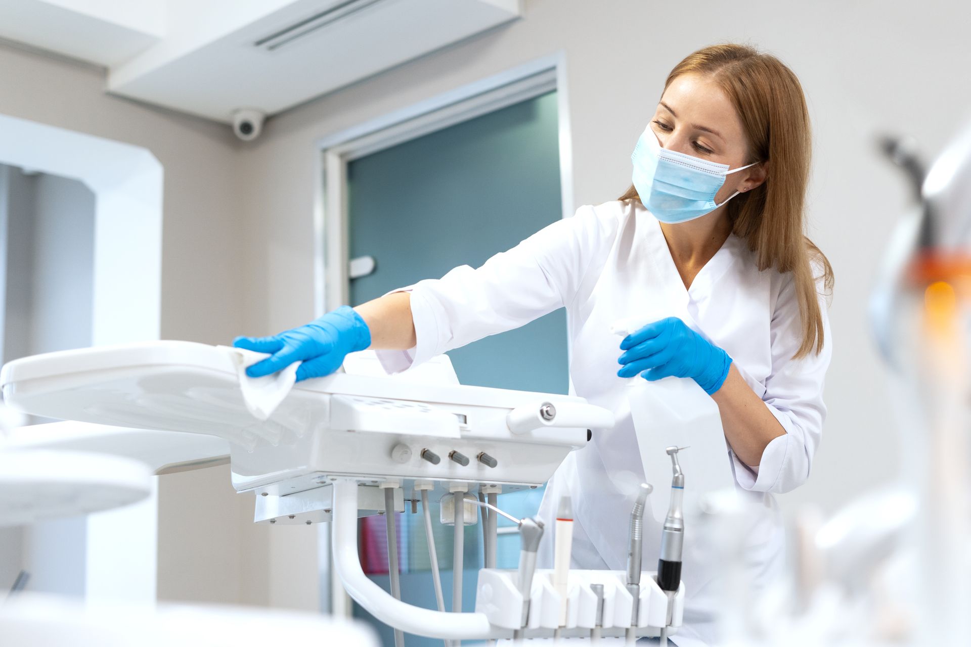 A person in a medical setting, wearing mask and gloves, cleaning dental equipment with wipes.