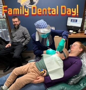 Family dental day: dentist examining child in mother's lap. Man in background, dental equipment visible.