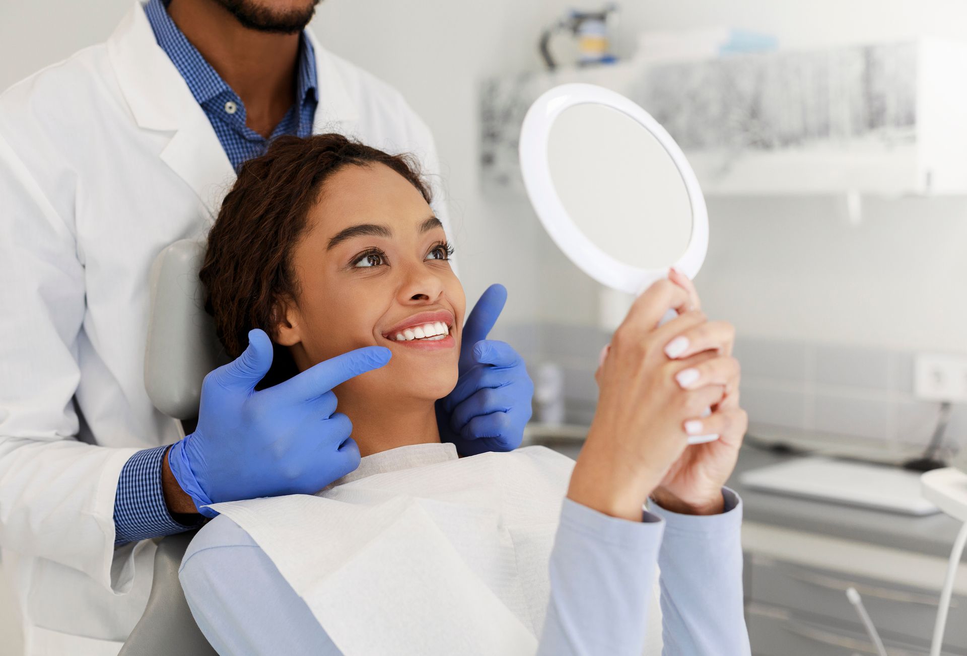 Dentist pointing at patient's teeth. Patient smiles, looking in a hand mirror. Clinic setting.