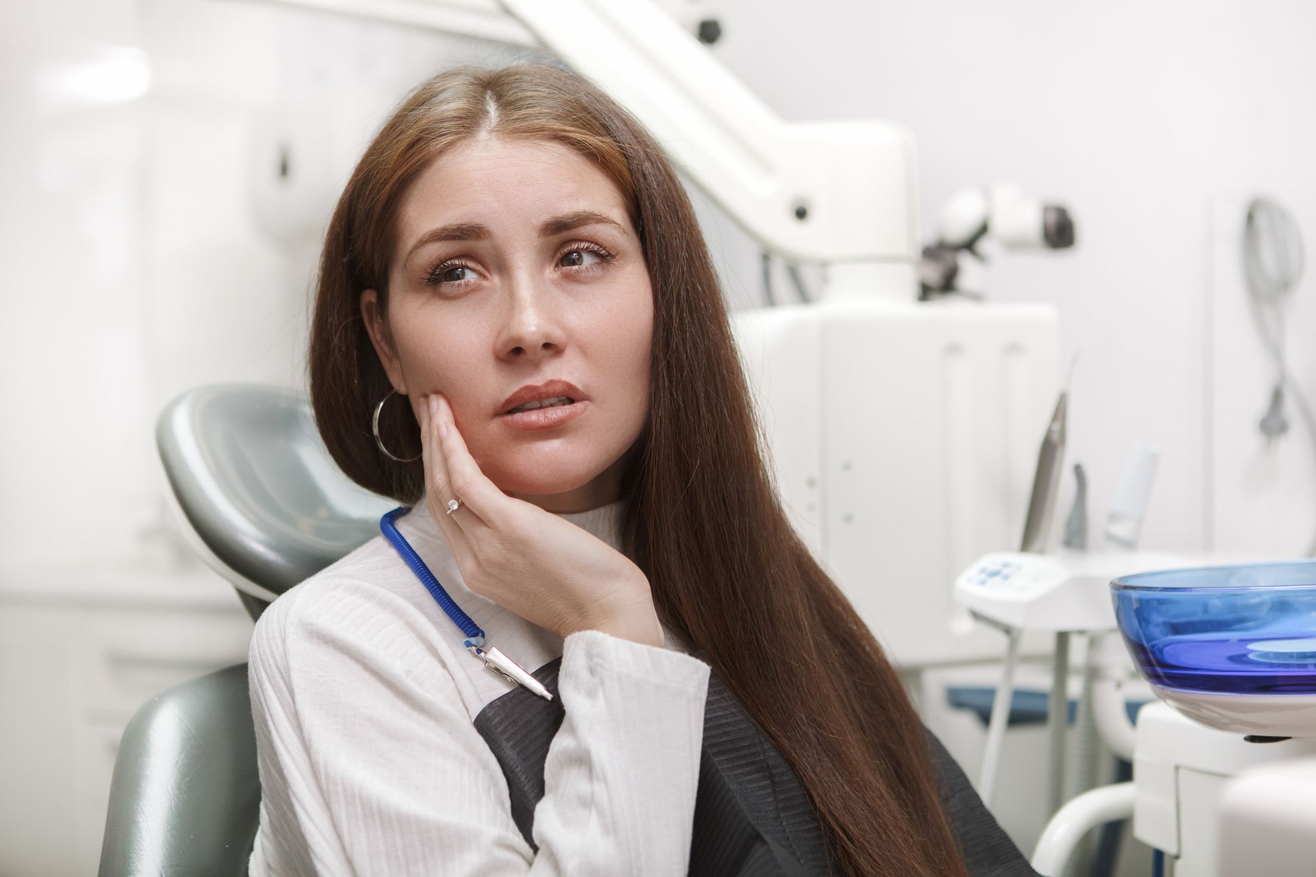 Woman in dentist chair holding cheek, looking distressed. Dental tools visible in the background.