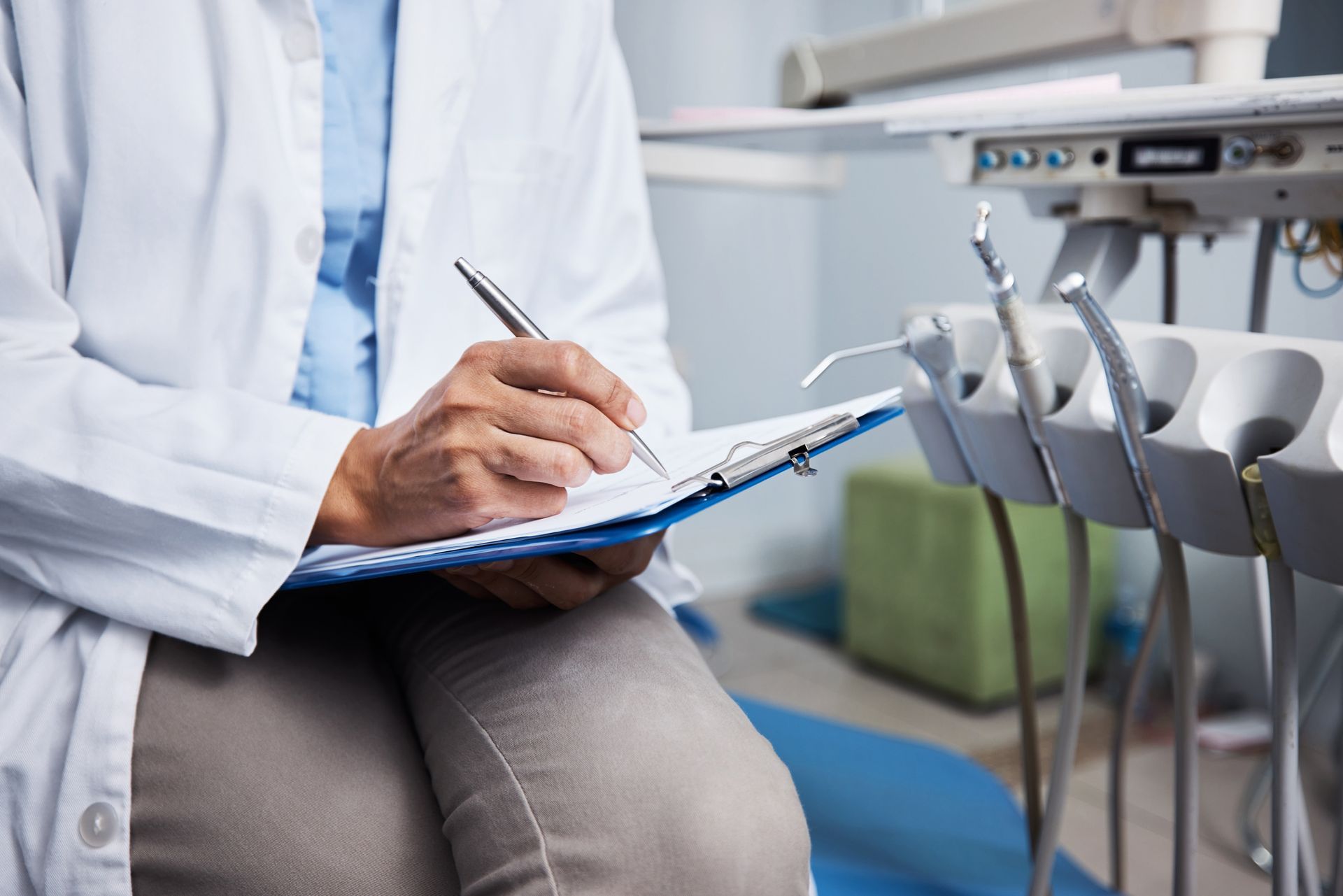 Dentist in white coat writing on clipboard next to dental instruments in an examination room.