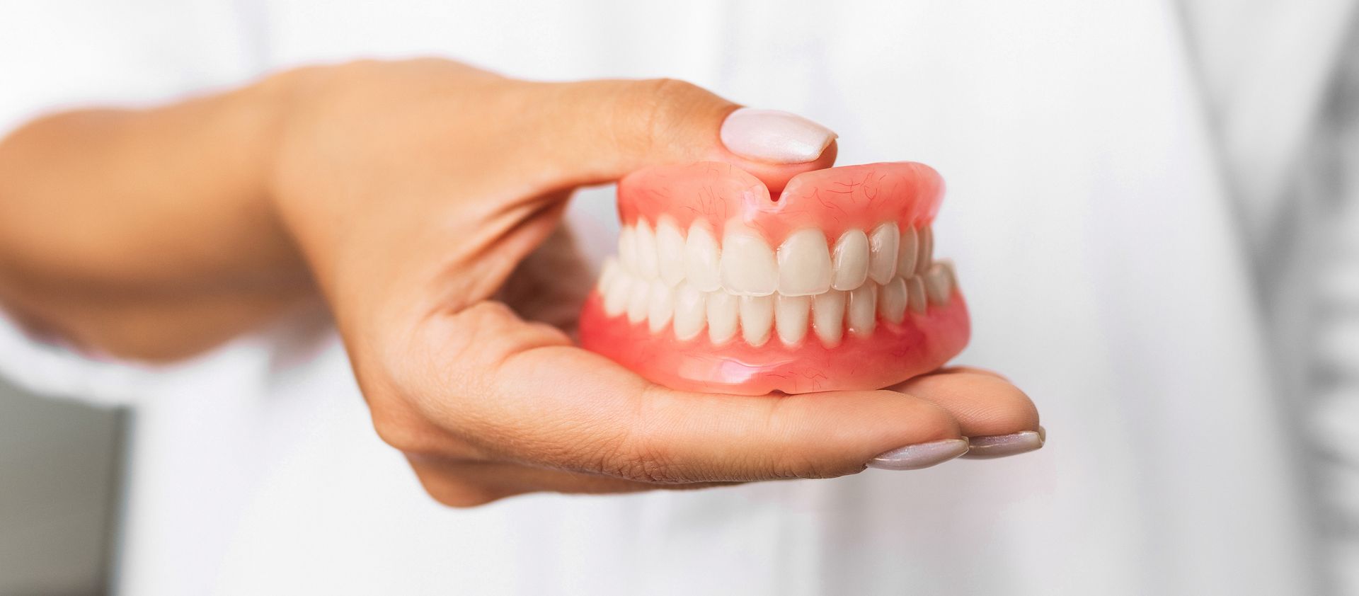 Person's hand holding a full set of dentures; the background is blurred and white.
