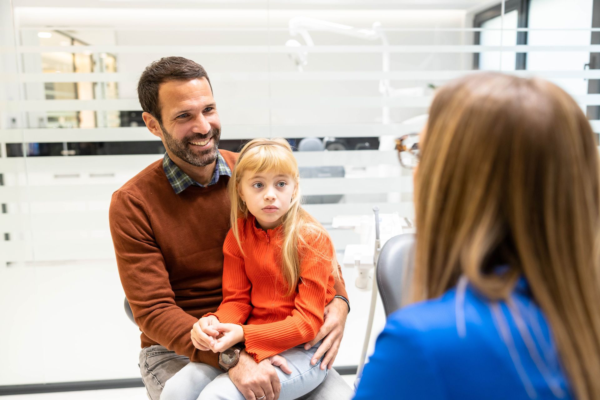 Father and daughter at dentist appointment. Dentist in blue speaks, daughter looks surprised, father smiles. Bright room.