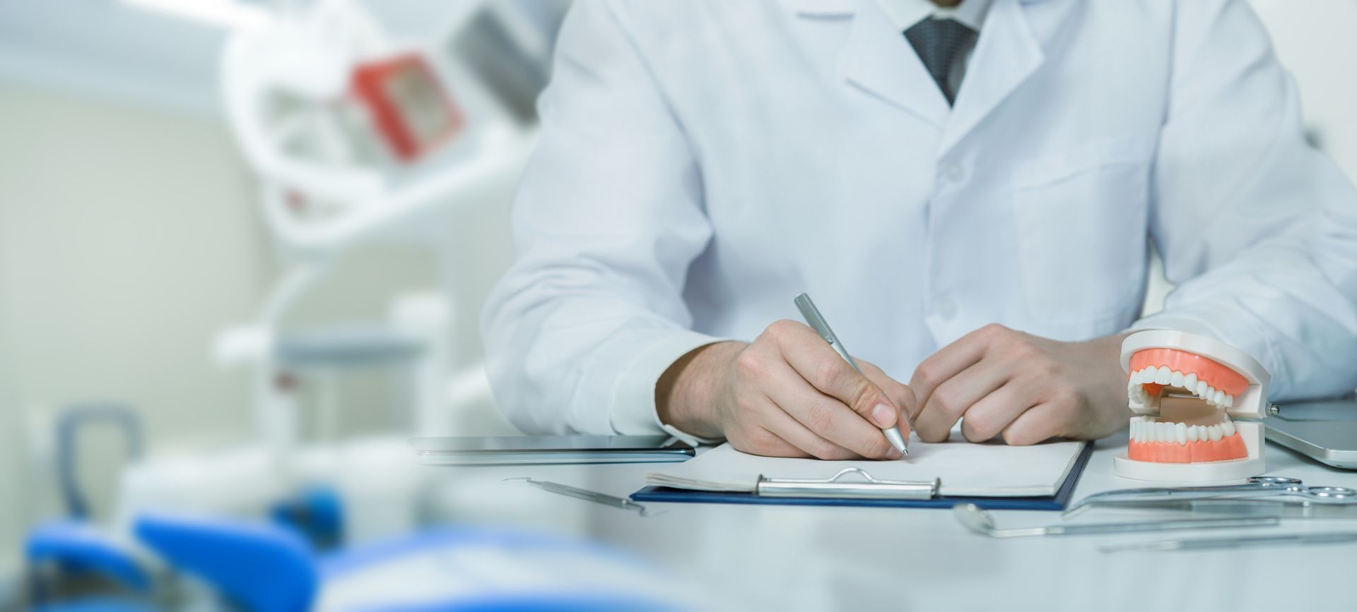 Dentist in white coat writing on a clipboard, dental model on the desk.