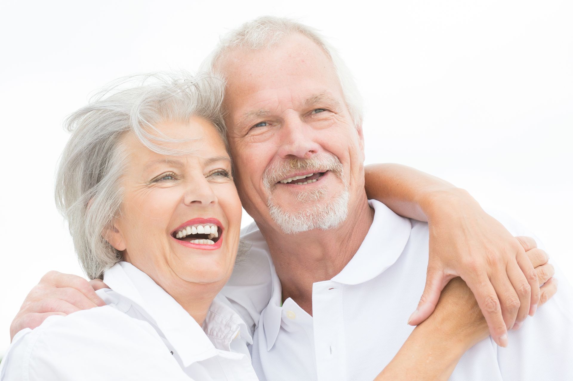 Smiling couple embracing, both with gray hair, wearing white shirts, outdoors.