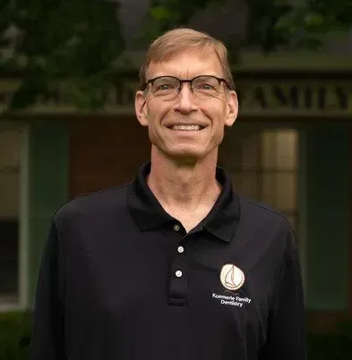 Man with glasses and black polo shirt smiling outdoors. Building in background.