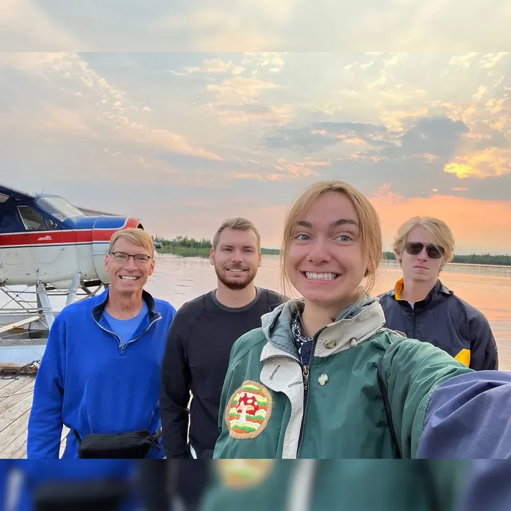 Four people stand in front of a seaplane on a dock at sunset, smiling.