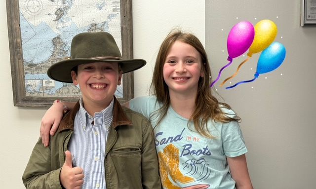 Boy in hat and jacket gives thumbs up, arm around girl in blue shirt; balloons float by.
