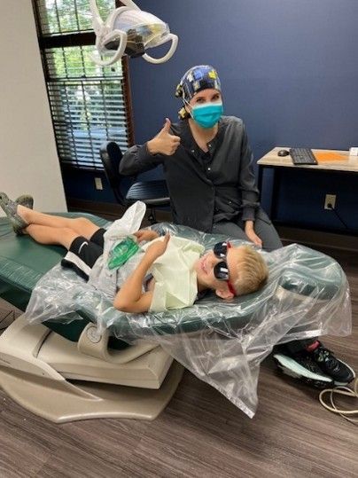 Girl in Santa hat and dental assistant in blue scrubs stand near Christmas tree.