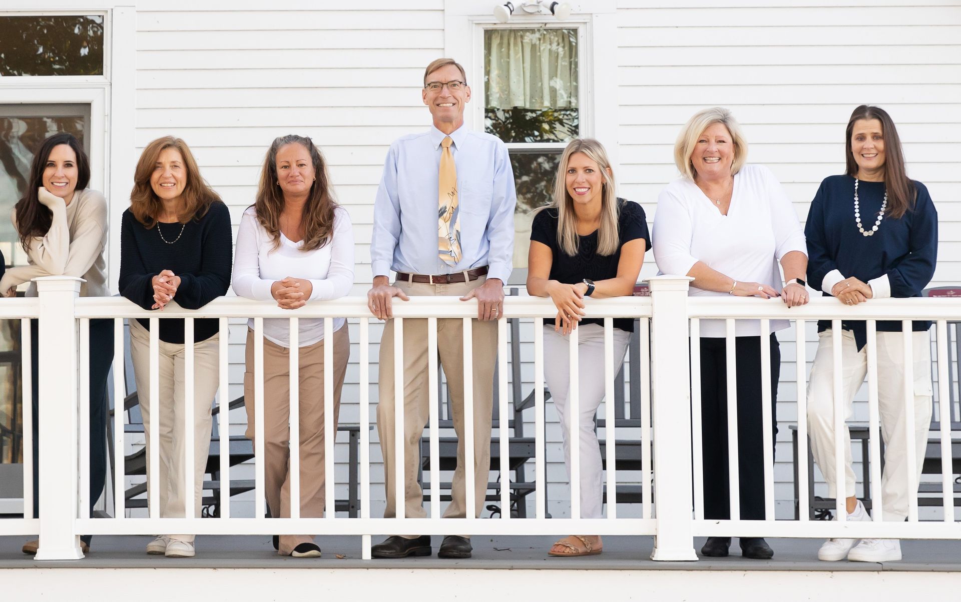 Group of seven people smiling, standing behind a white porch railing in front of a white house.