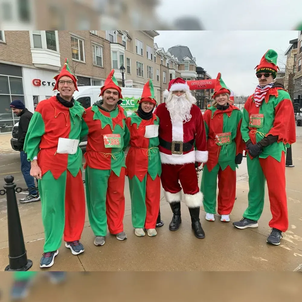 Santa Claus with elves in red and green costumes on a street with buildings.