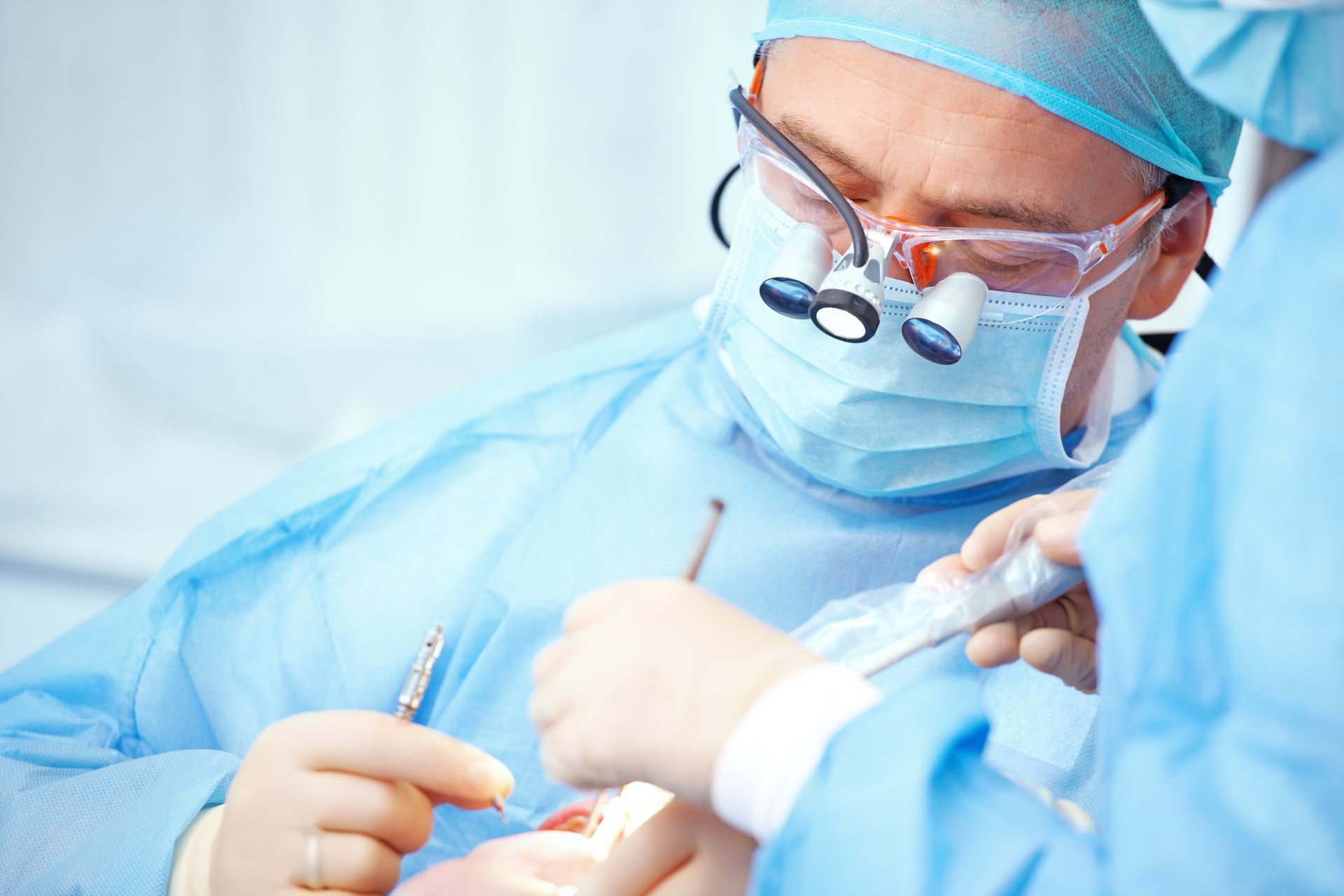 Surgeon in blue scrubs, mask, and glasses operating with surgical tools; focused expression.