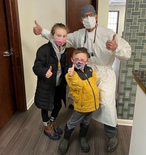 Two children and a dentist in protective gear give thumbs up in a hallway.