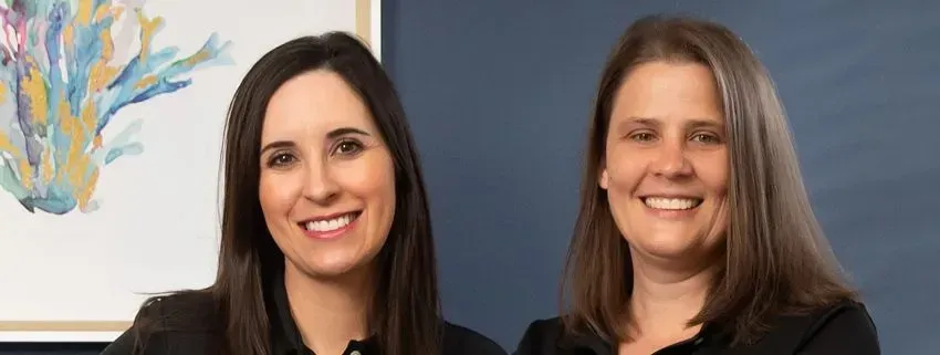 Two women in black polo shirts smile in front of a painting. One has hands on her hips, the other has arms crossed.