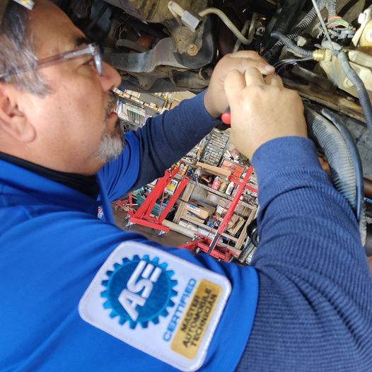 Technician in blue uniform inspecting machinery with tools in a tight industrial space | Auto Pros