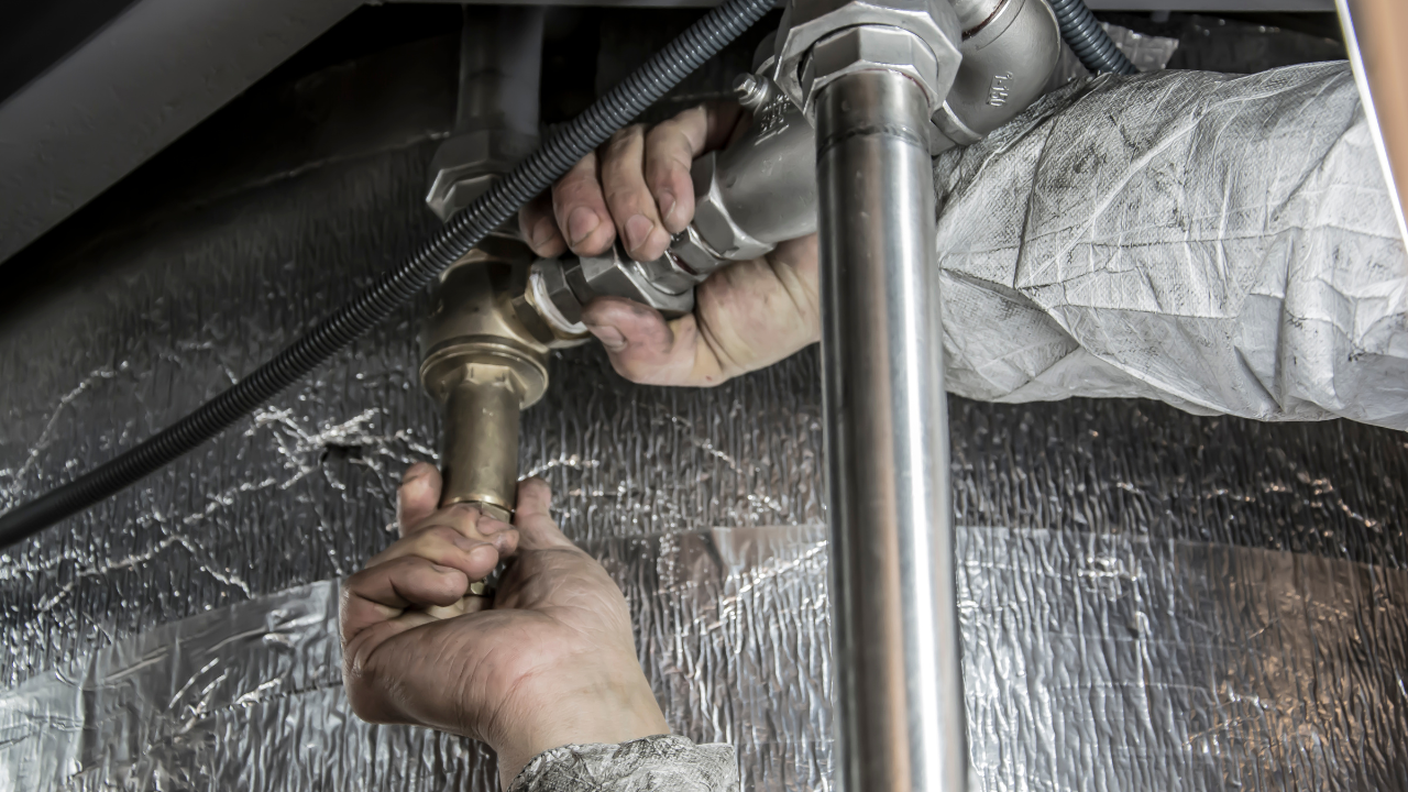 A worker wearing protective sleeves performs maintenance on silver metal pipes in a dark, foil-lined space.