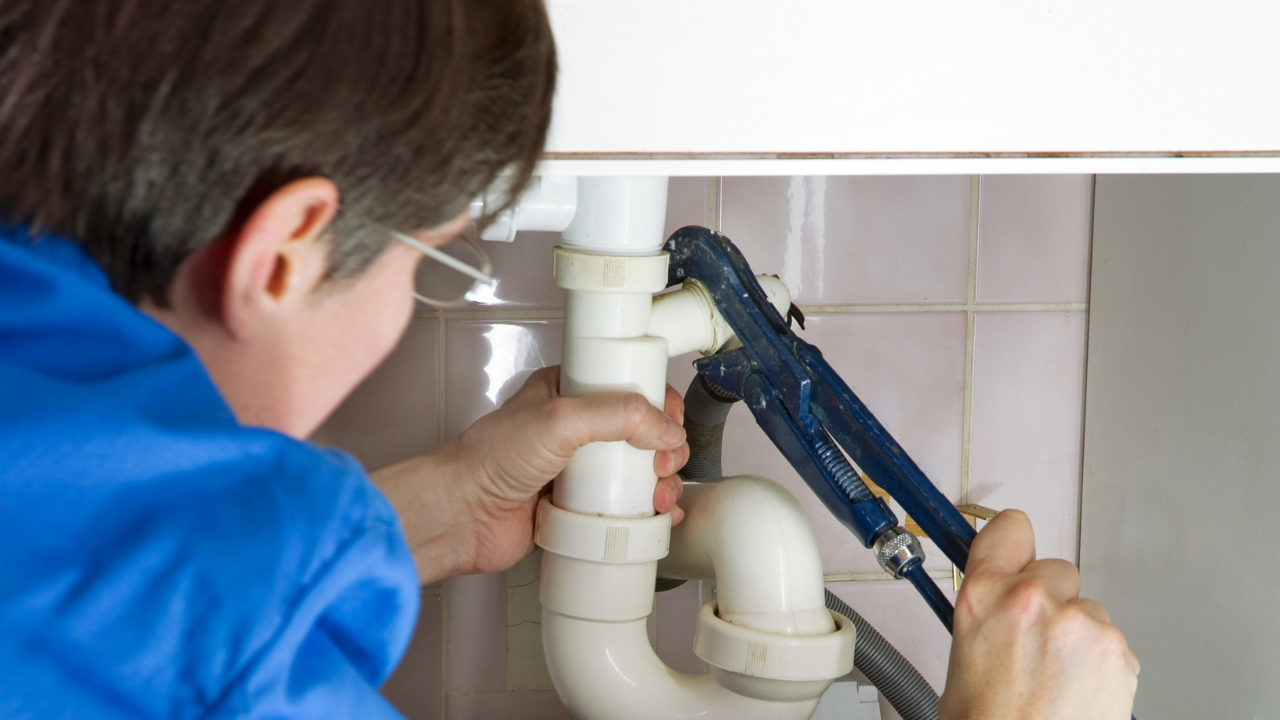 A person in a blue shirt uses a blue pipe wrench to tighten plastic plumbing pipes under a sink.