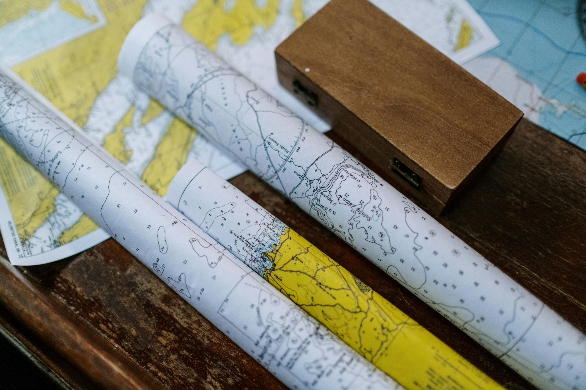 Three nautical charts are rolled up and resting on a wooden surface next to a rectangular wooden box.