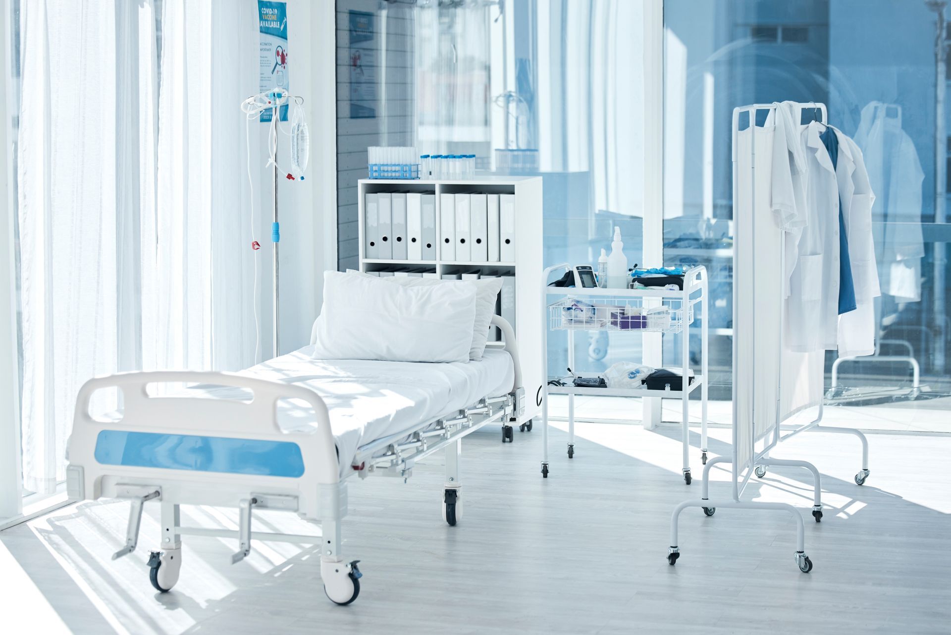 Bright hospital room with empty bed, medical cart, and hanging white coats in a clean, sunlit setting