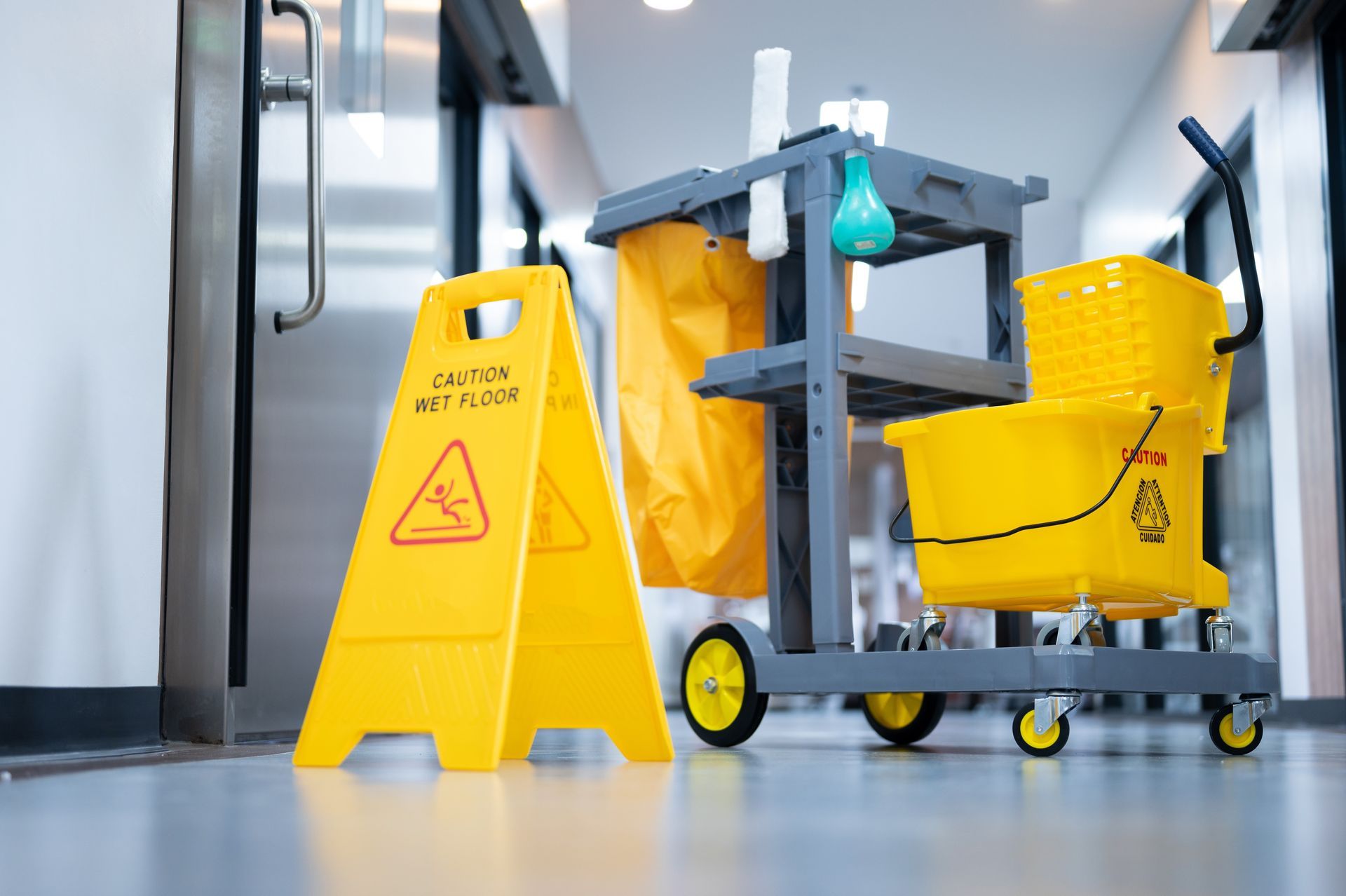 Yellow cleaning cart and caution sign in a bright hallway with a mop bucket and supplies.
