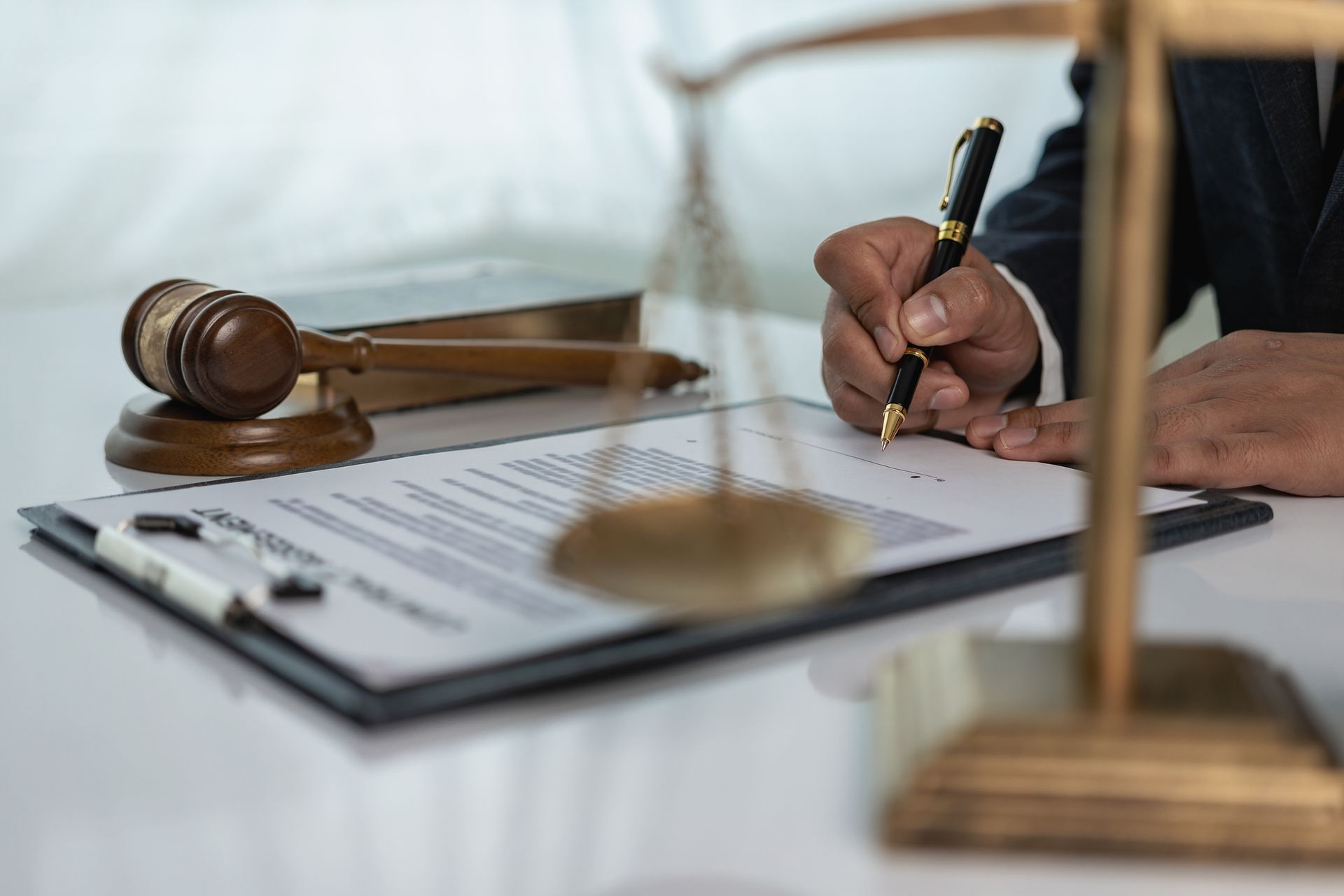 Person signing legal document with gavel and scales on desk.