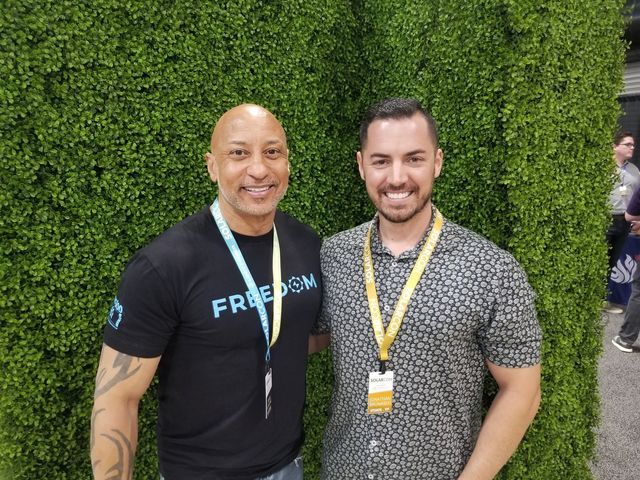 Two men are posing for a picture in front of a green wall.
