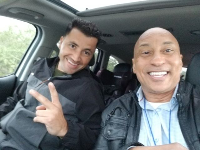Two men are sitting in a car and one is giving a peace sign