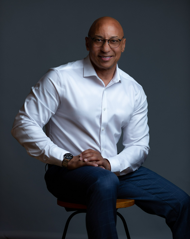 A man in a white shirt and blue jeans is sitting on a stool.