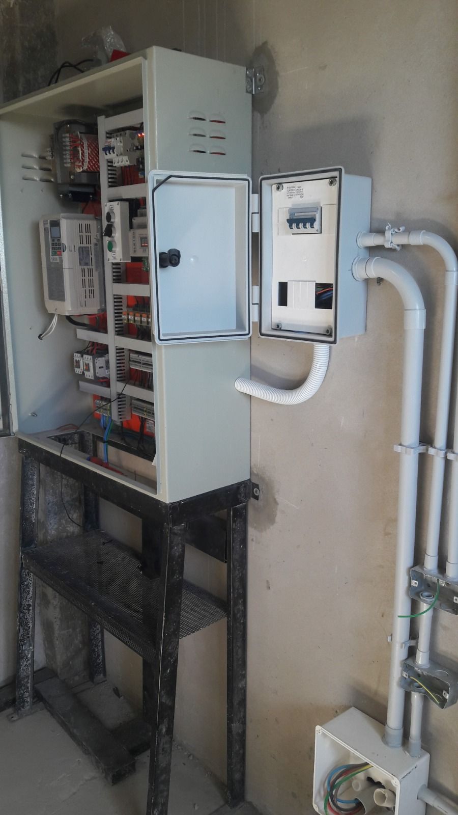 Panel de control eléctrico montado sobre un soporte, con puerta abierta que revela componentes internos y conductos para cableado.