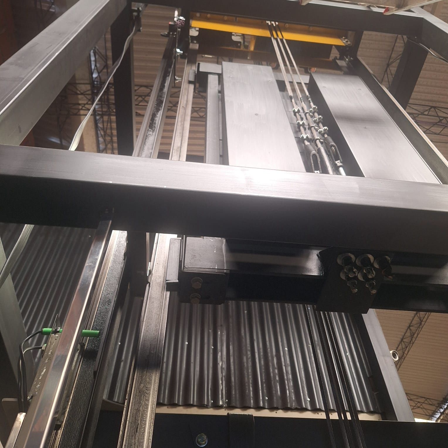 Mirando hacia arriba al mecanismo de un ascensor en un gran entorno industrial, con cables de acero y rieles visibles.