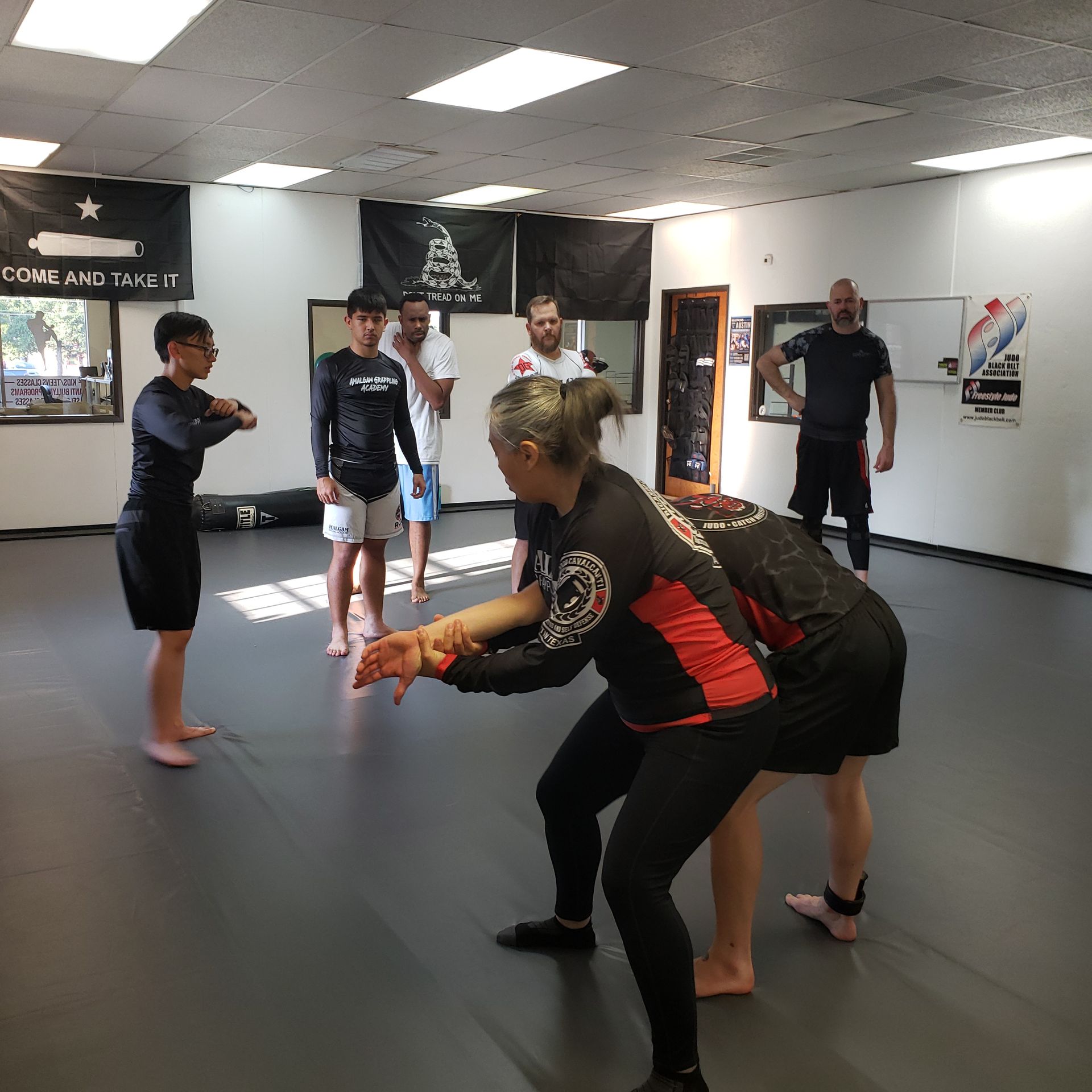 People training jiu-jitsu in a gym. A woman demonstrates a move while others watch. Black mats, flags.