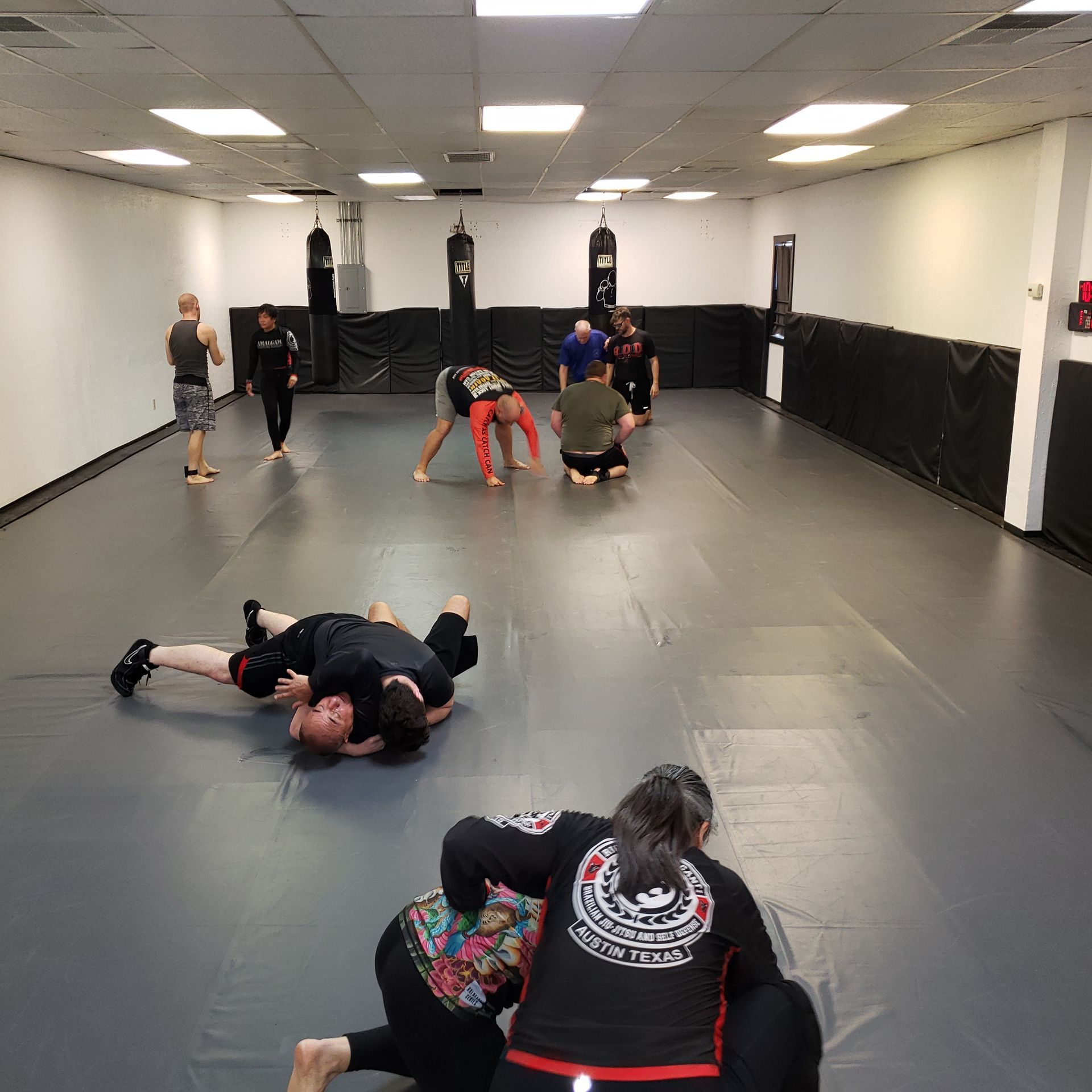 People practicing martial arts on a large mat in a training room. Several are in combat positions.