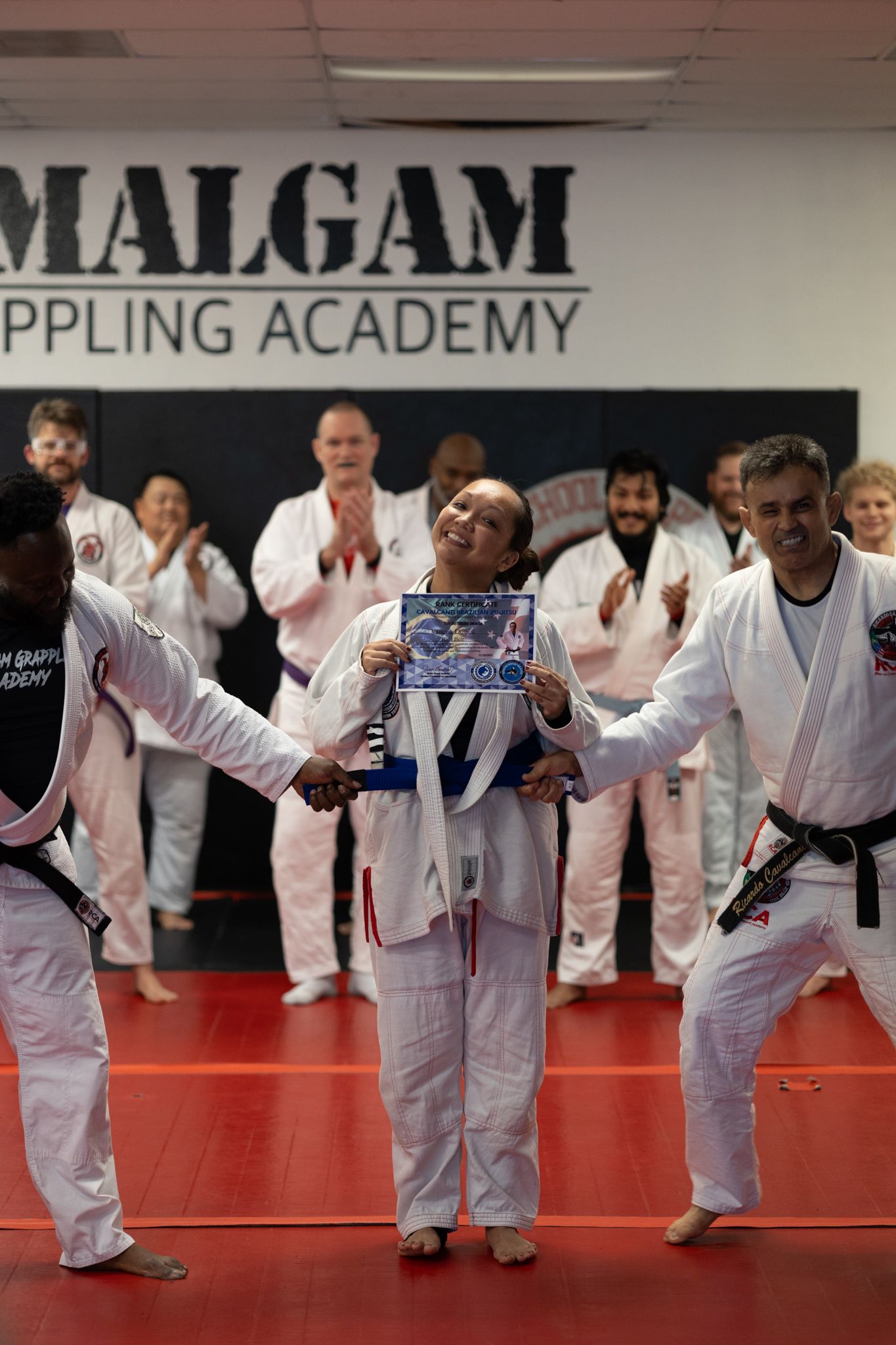 People in white uniforms at a grappling academy, celebrate a person in a blue belt holding a framed photo.