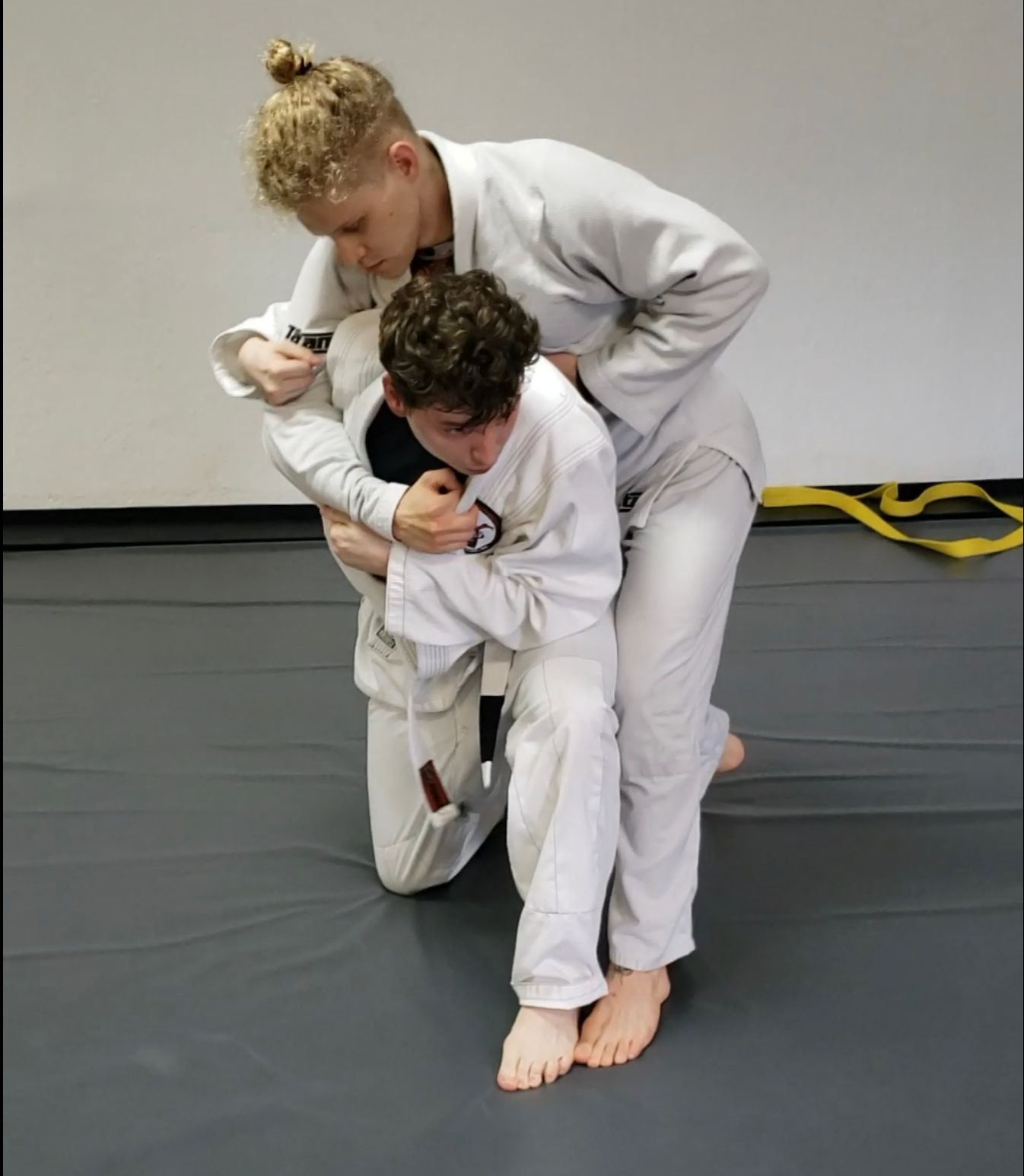 Person in white gi kneels, being lifted by another in white gi during a martial arts class.