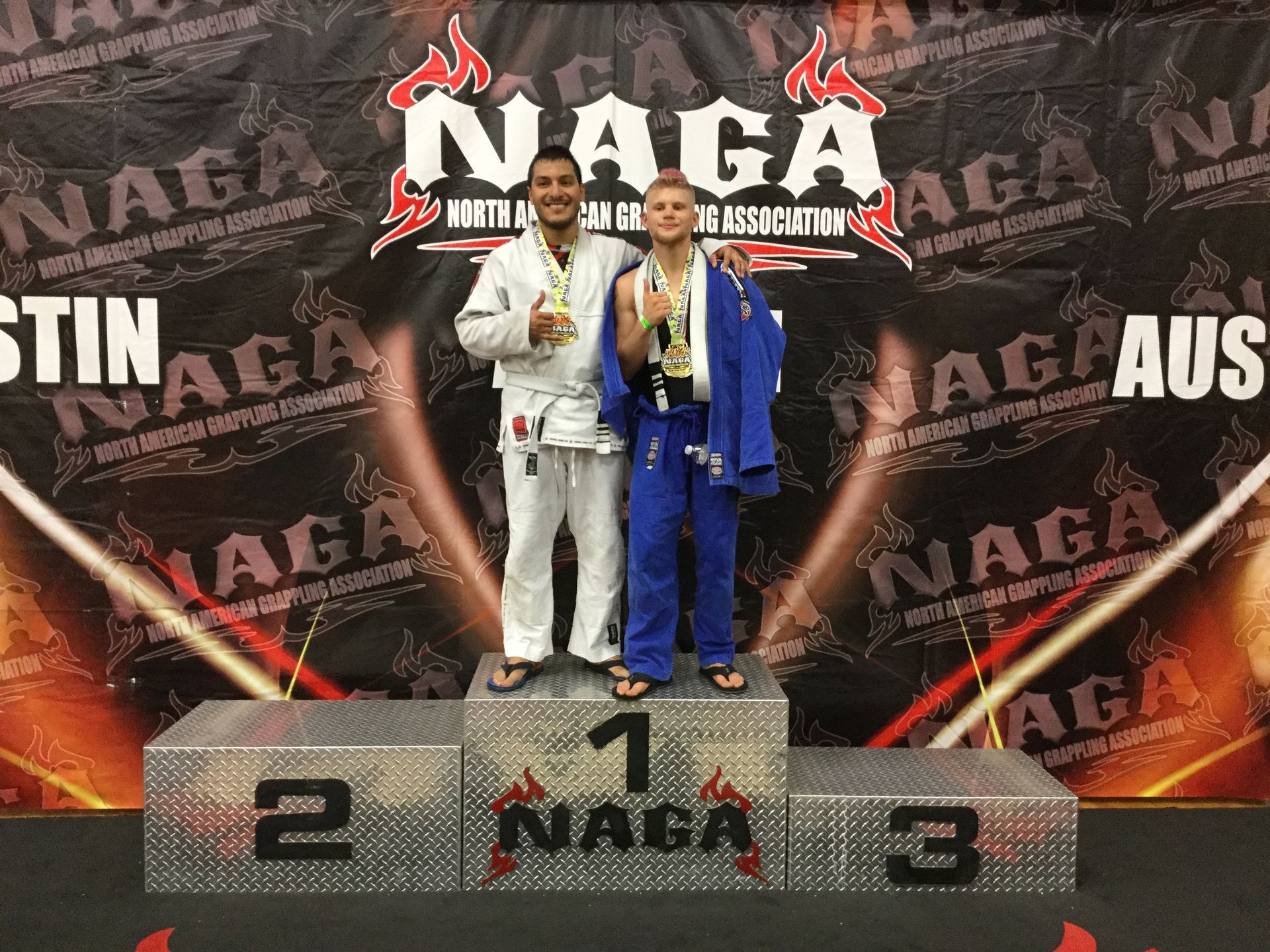 Two men on a podium at a NAGA competition, wearing medals. The first place winner is in a blue gi.