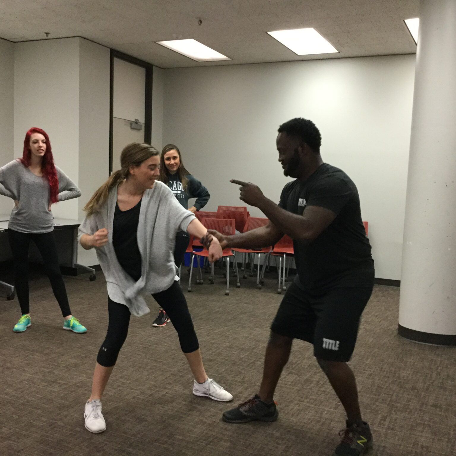 Woman and man practicing self-defense in a room. Man points and woman punches; two others watch.