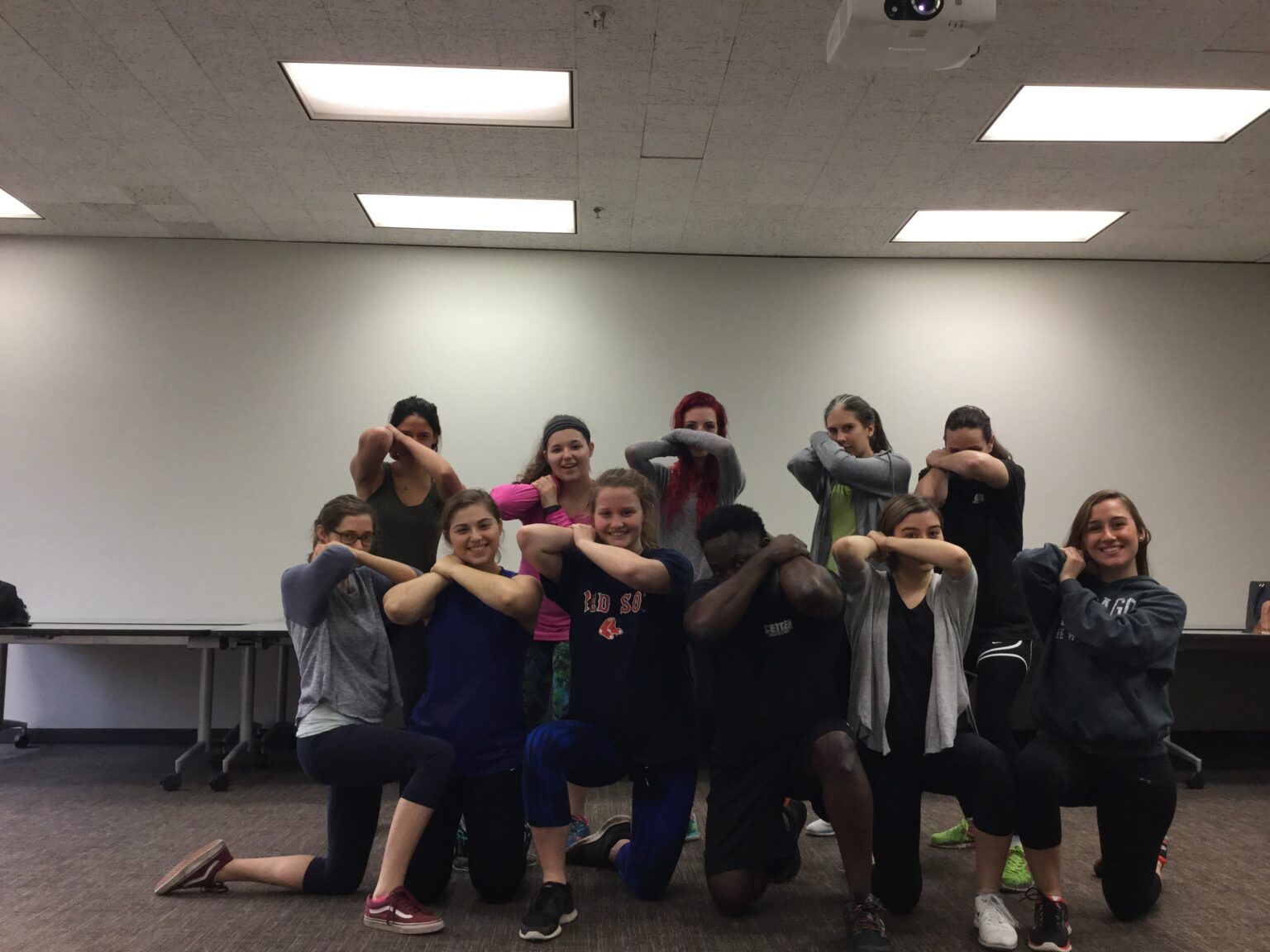 Group of teens, indoors, posing with arms crossed in front of their faces, in a dance-like position.