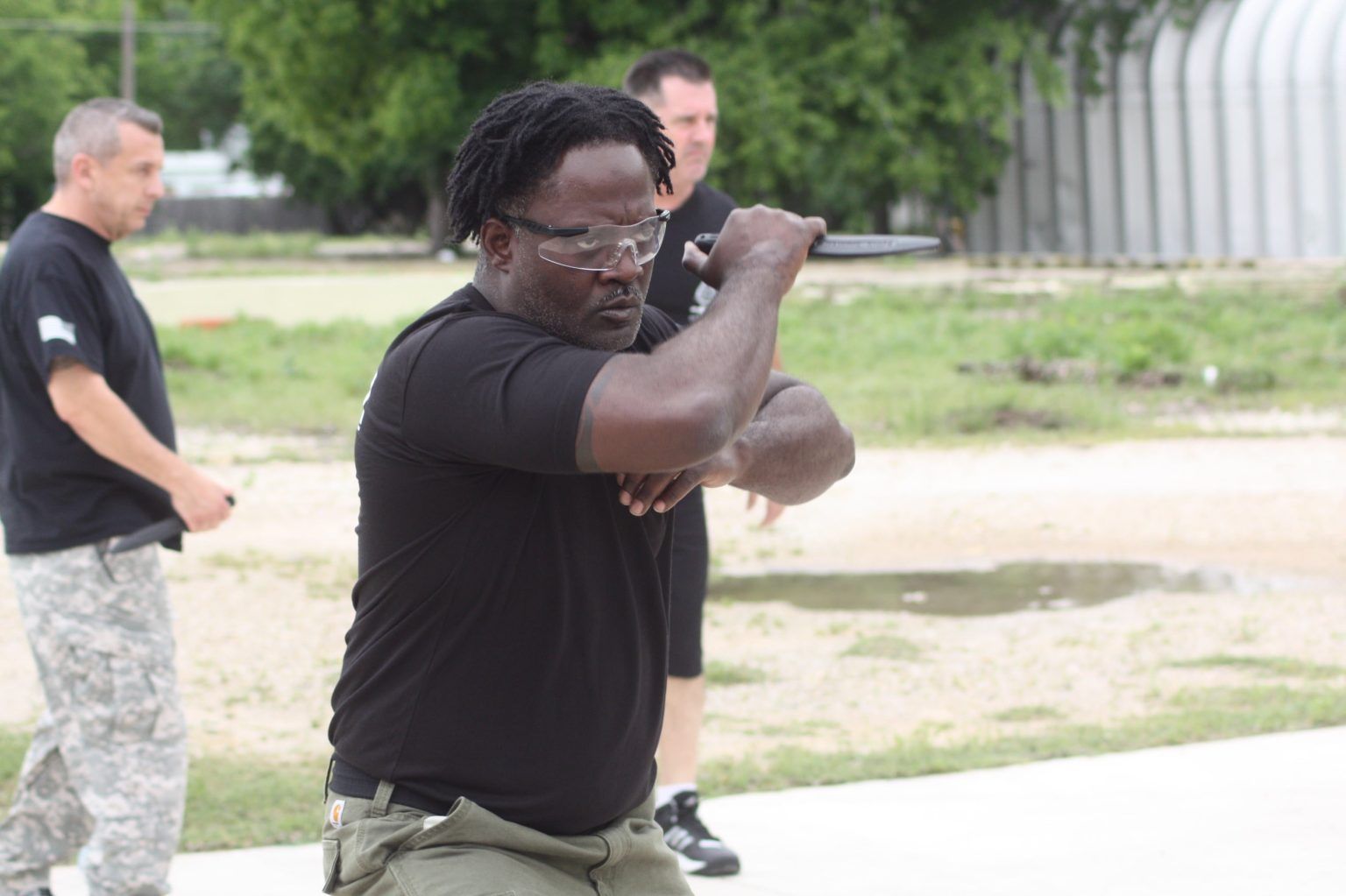 Man in safety glasses practices knife defense with two other men nearby.