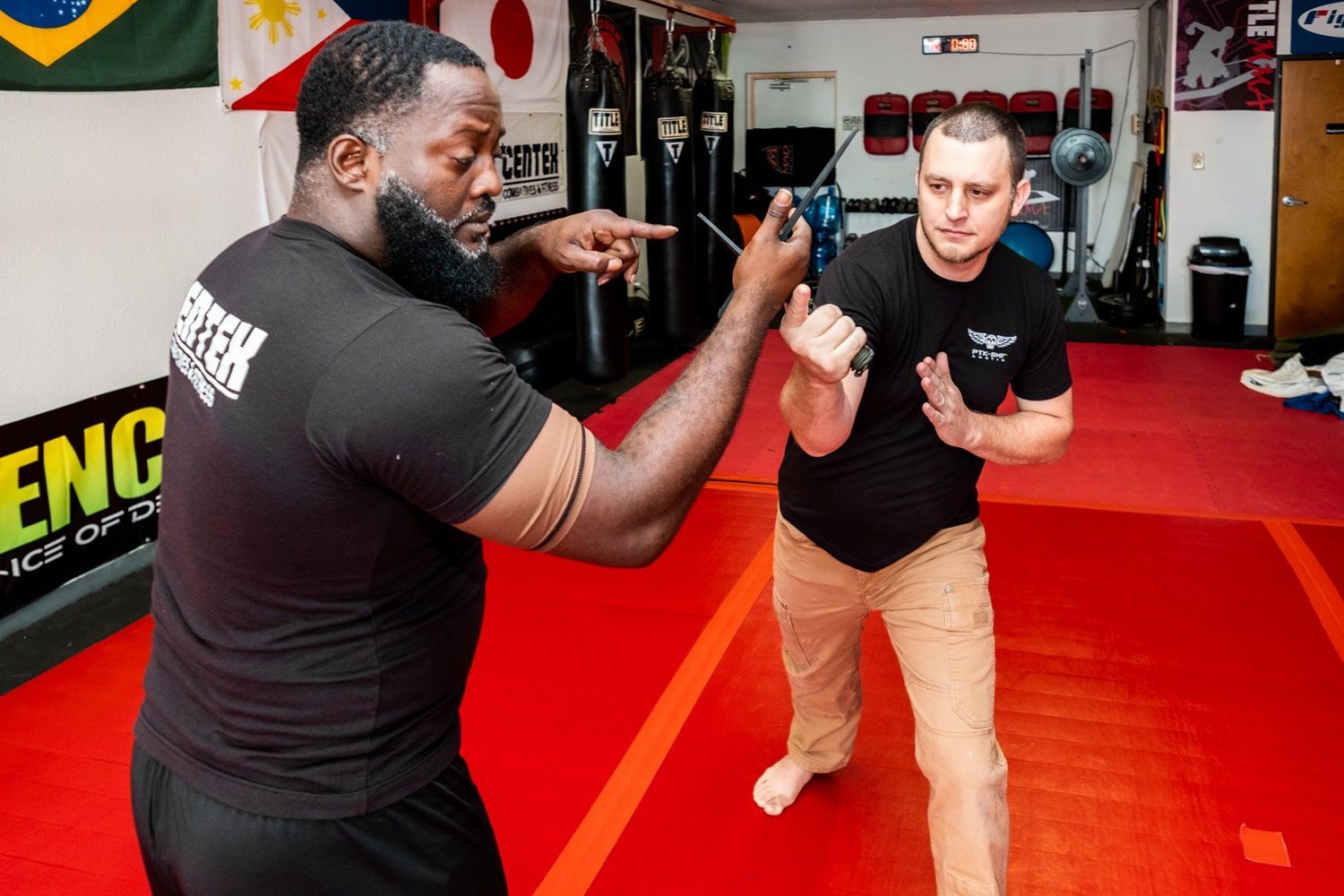 Two men practicing self-defense in a dojo. One points a finger, the other blocks with hands up. Red mat.