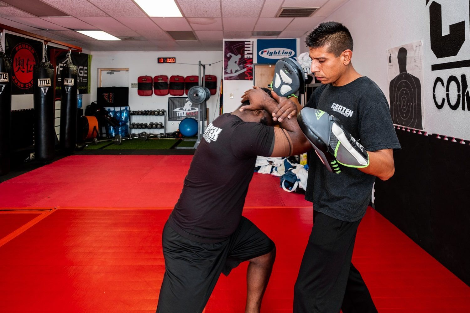 Man holding pads as another man practices boxing, indoors, on a red mat.