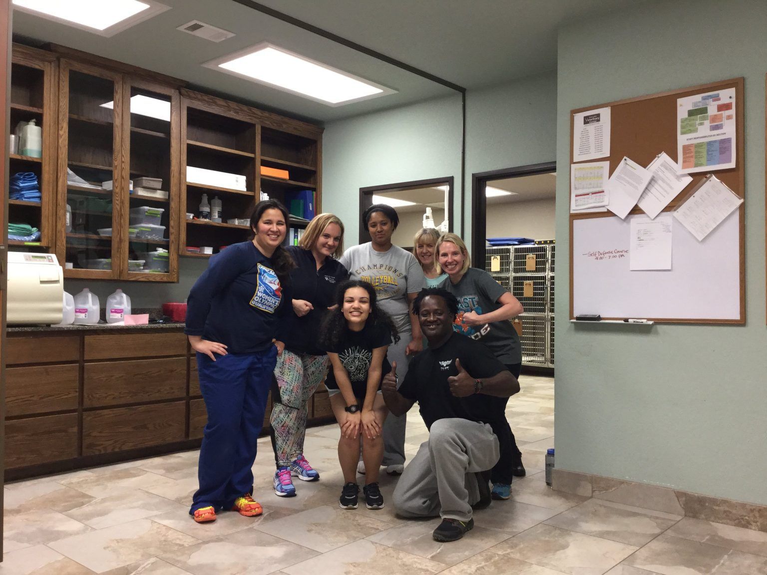 Group of seven people smiling in a medical office, posing in front of cabinets and a corkboard.