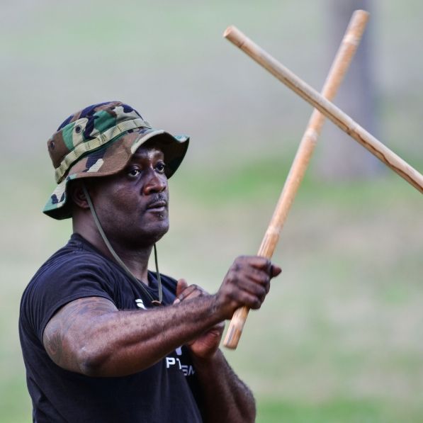 Man in camo hat, holding two wooden sticks in an outdoor training session, practicing a martial art.