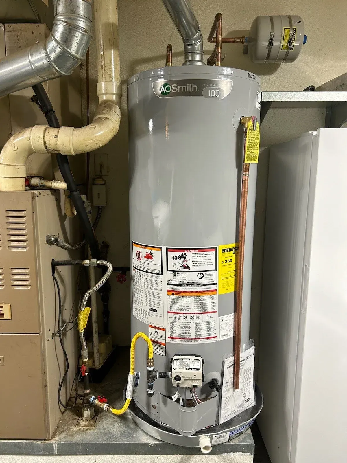 A tall, silver water heater stands next to a furnace in a basement setting. Yellow gas line visible.