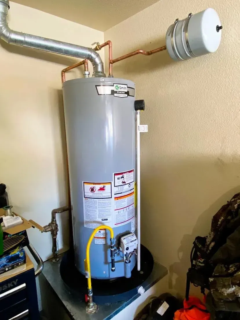 Tall gray water heater with copper pipes, a gas line, and an expansion tank, set in a garage corner.