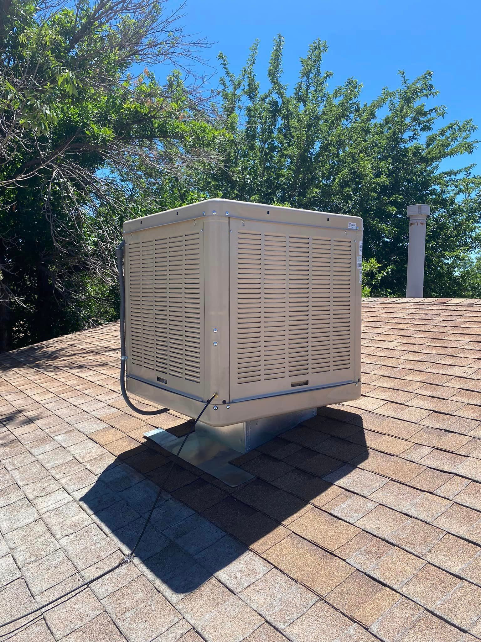 Cooling unit on a rooftop, tan and square, with dark shadow under clear blue sky and trees.