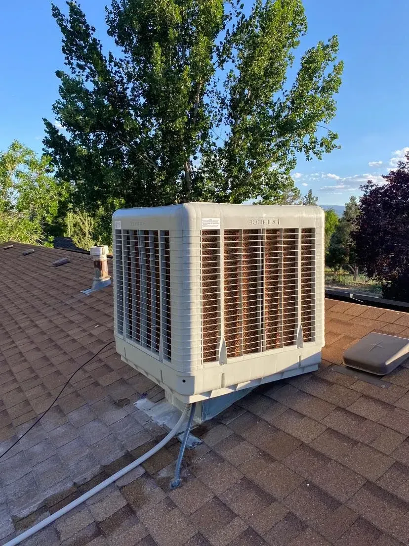 Evaporative cooler on a brown shingled roof, with a blue sky and tree in the background.