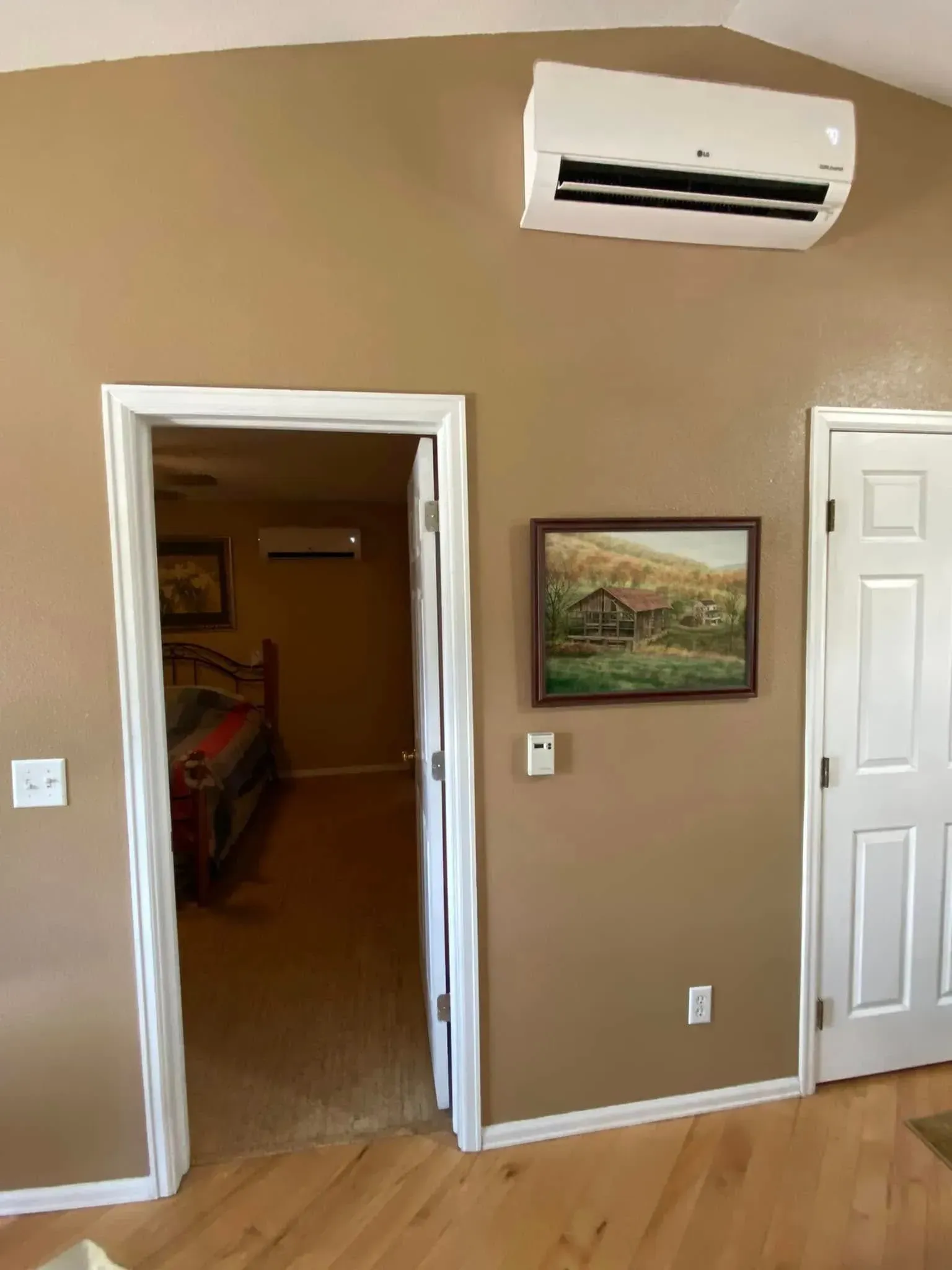 A room with tan walls, white doorframes, and an air conditioning unit. A painting and a doorway are visible.